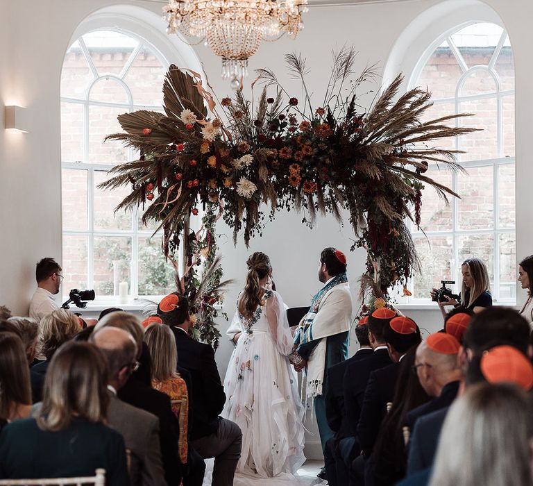 Bride in long mesh sleeve wedding dress with floral embroidery stands with groom in kippah and green suit under arch with dried flower cloud for interfaith wedding ceremony