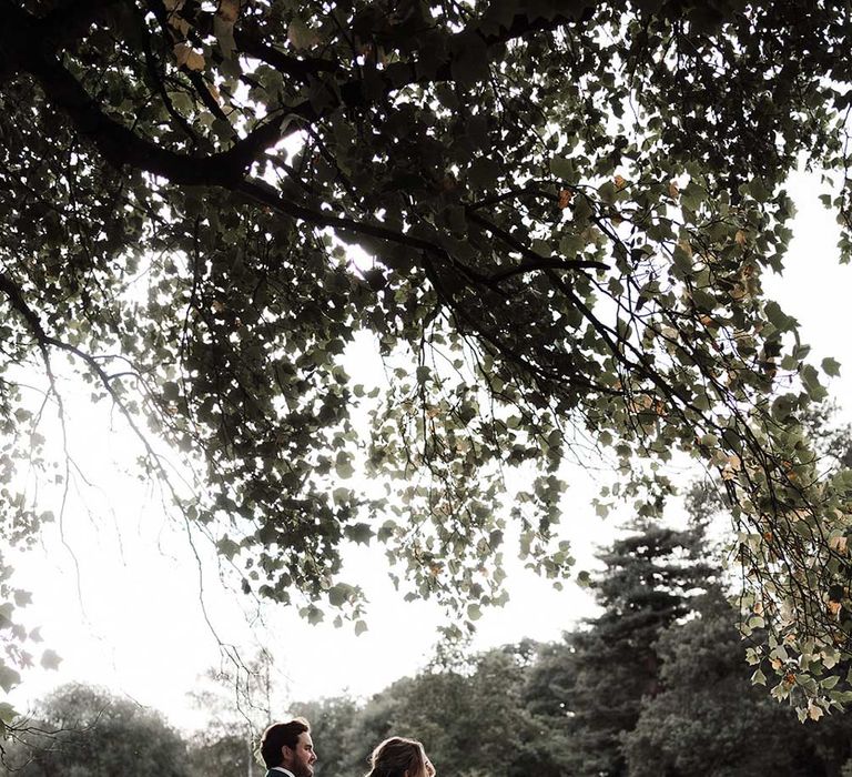 Groom in blue suit walks with bride in low back wedding dress with train through grounds of Iscoyd Park