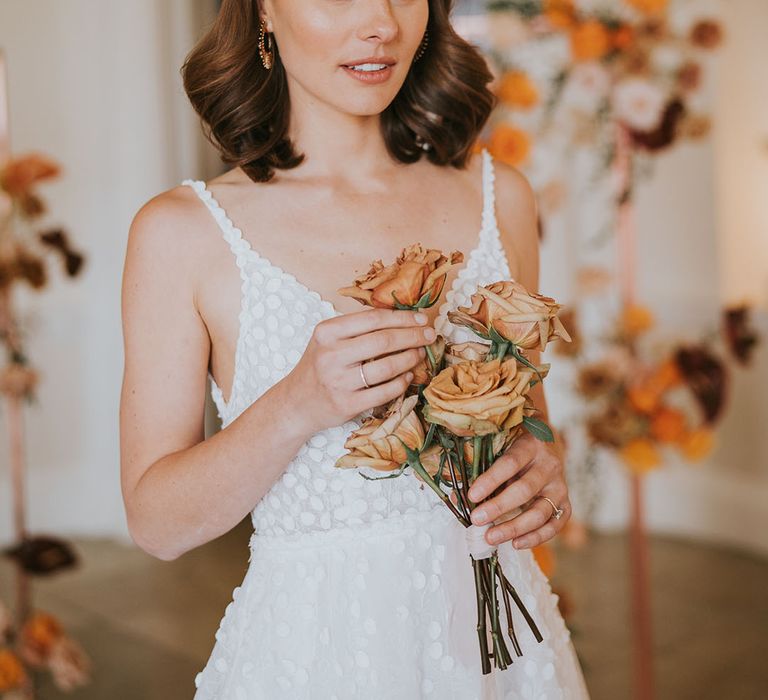 Bride with shoulder length hair in a pearl headband holding orange peeled back rose stems 