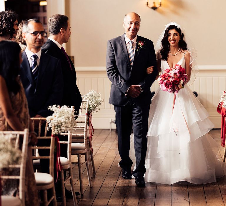 Bride in white layered tulle wedding dress with plunging neckline and thin straps wearing veil walks down the aisle at Elmore Court with man in dark suit and striped tie