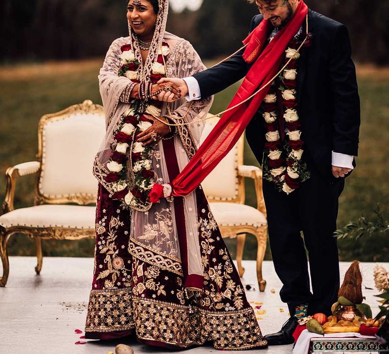 Bride wears Indian burgundy saree wedding dress and holds hands with groom who wears matching tie 