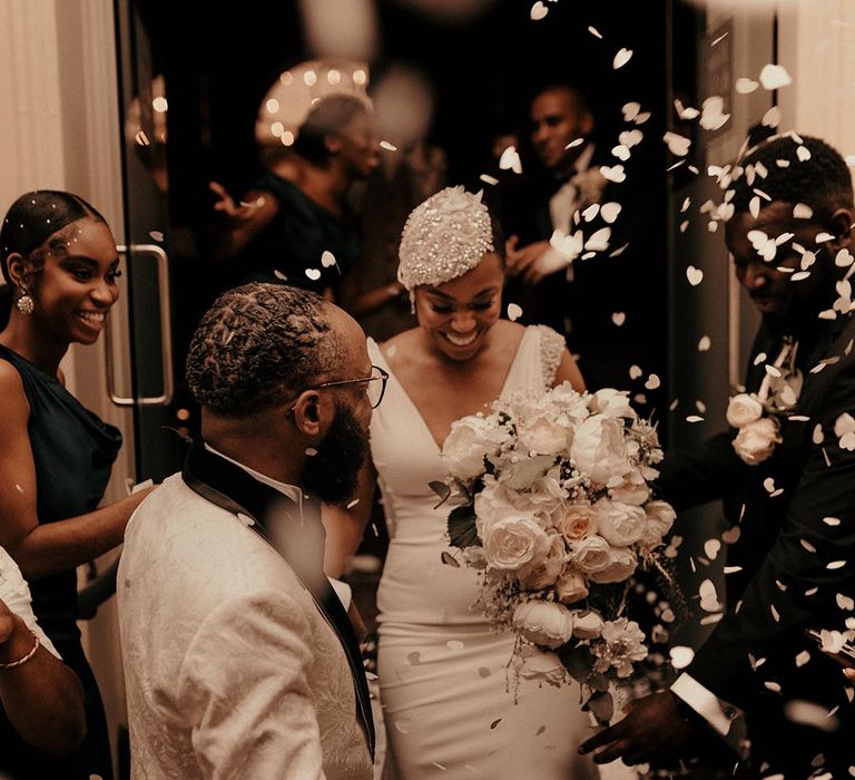 Confetti moment with groom in a white brocade tuxedo jacket helping his bride in a fitted wedding dress with beaded headdress holding a white flower bouquet 