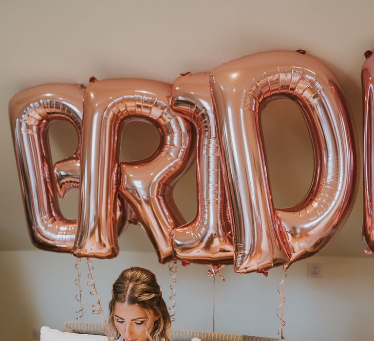 Bride in satin dressing gown reads letter and opens gift from the groom in front of rose gold 'bride' balloons