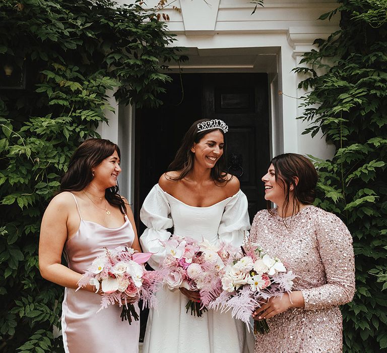 Bride in off the shoulder wedding gown with tiara crown stands with bridesmaids with one in pink satin dress and the other in pink high neck sparkly dress