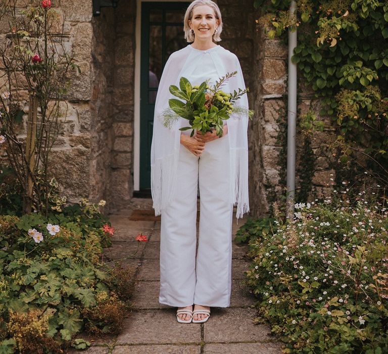 Bride in handmade bridal jumpsuit with cape and white block heels and short hair with silver jewellery and green leaf wedding bouquet