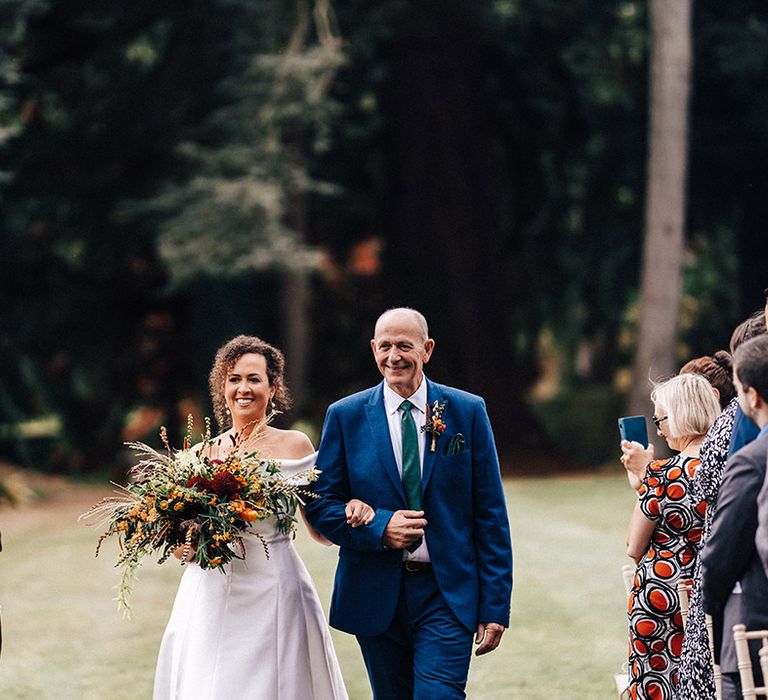 Bride in off the shoulder wedding dress carrying autumnal bouquet is walked down the aisle by father in blue suit