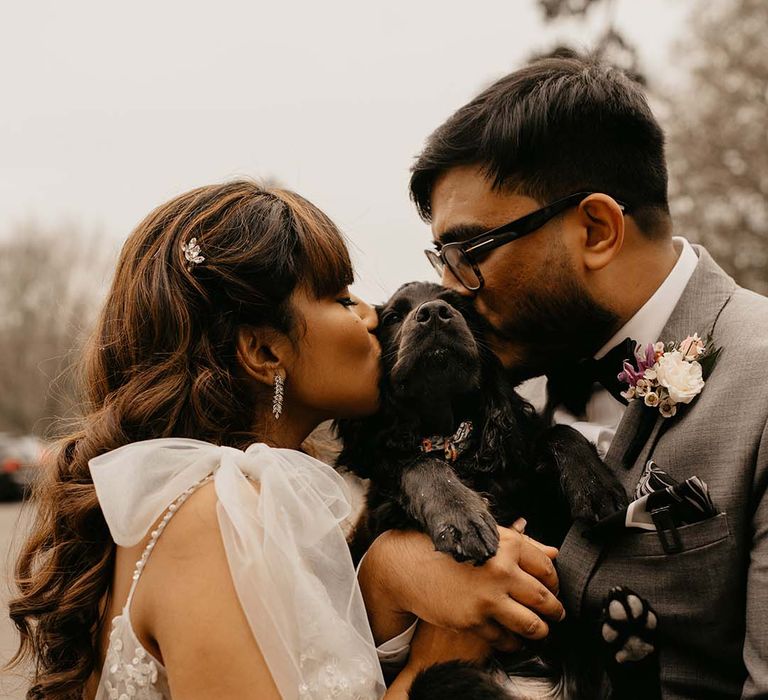 Bride in white wedding dress with silver hair accessories and earrings holding black puppy with groom in grey suit