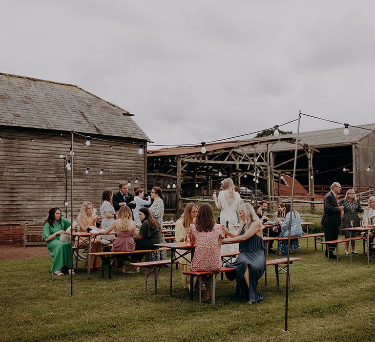 Wedding guests socialising and drinking on red benches outside the rustic barn venue 