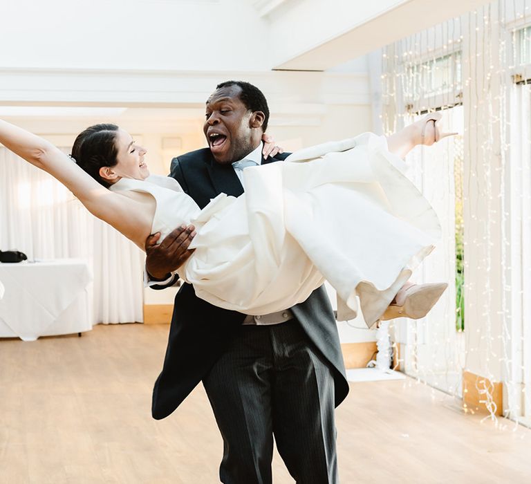 Groom in black morning suit lifts the bride in a princess carry as they dance together 