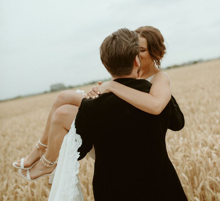 Groom lifts the bride and kisses her as they stand in a field together |