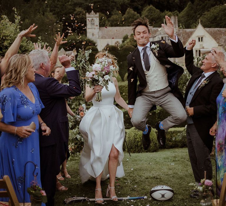 Groom jumps for joy over a rugby ball with bride as guests throw confetti in congratulations 