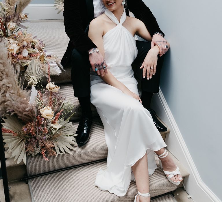 Groom in a tuxedo sitting on the stairs with his bride in a satin halter neck wedding dress surrounded by preserved pastel flowers and dried grasses 