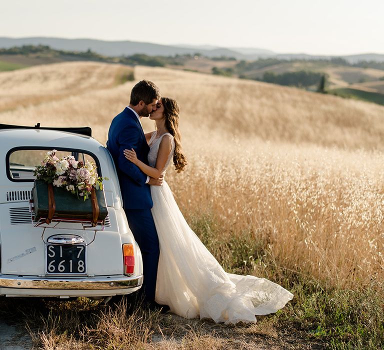 Bride & groom stand beside vintage Fiat 500 in golden fields in Tuscan countryside on their wedding day