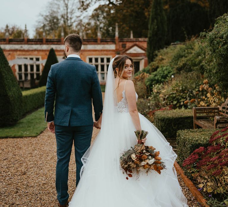 Bride wearing a layered tulle wedding skirt and sequinned top wedding dress looks back at the camera walking with the groom in a blue suit 