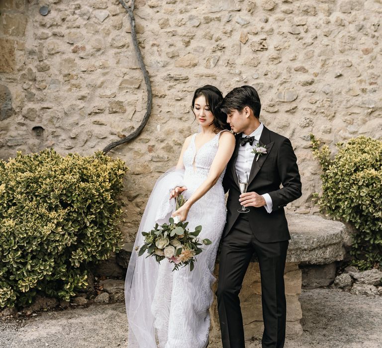 Groom in black-tie holds champagne glass and stands beside his bride who wears her dark hair in loose curled bun to the side