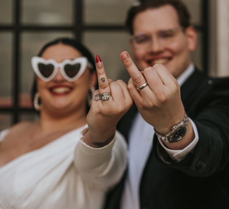 Bride wears heart-shaped sunglasses as she and her groom hold up their wedding fingers to the camera