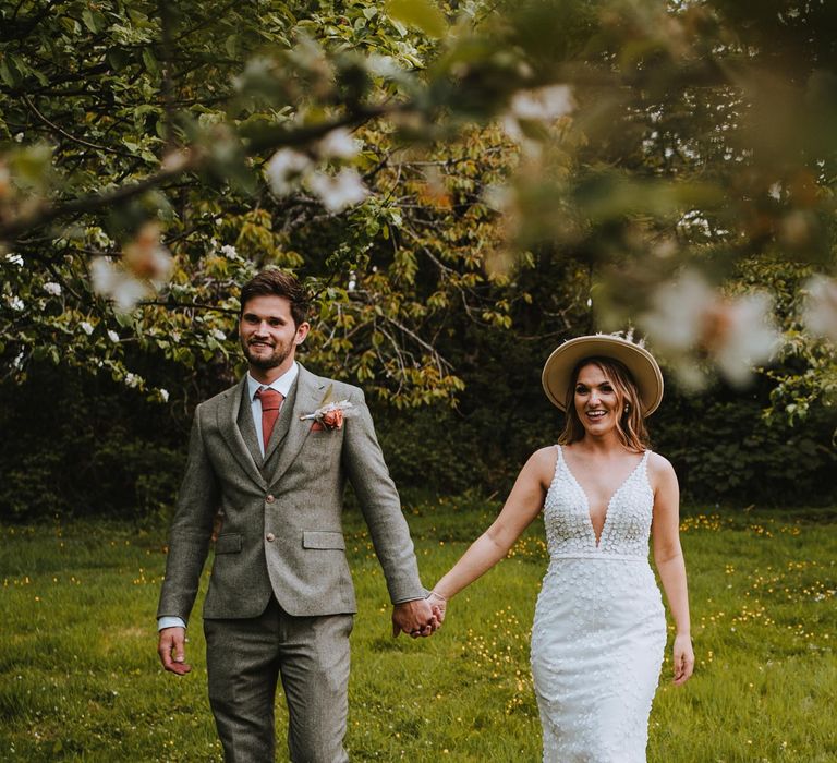 Bride in sleeveless Made With Love Bridal dress with white embellishments and groom in grey suit with orange tie, pocket square, and boutonniere holding hands at The Green Cornwall