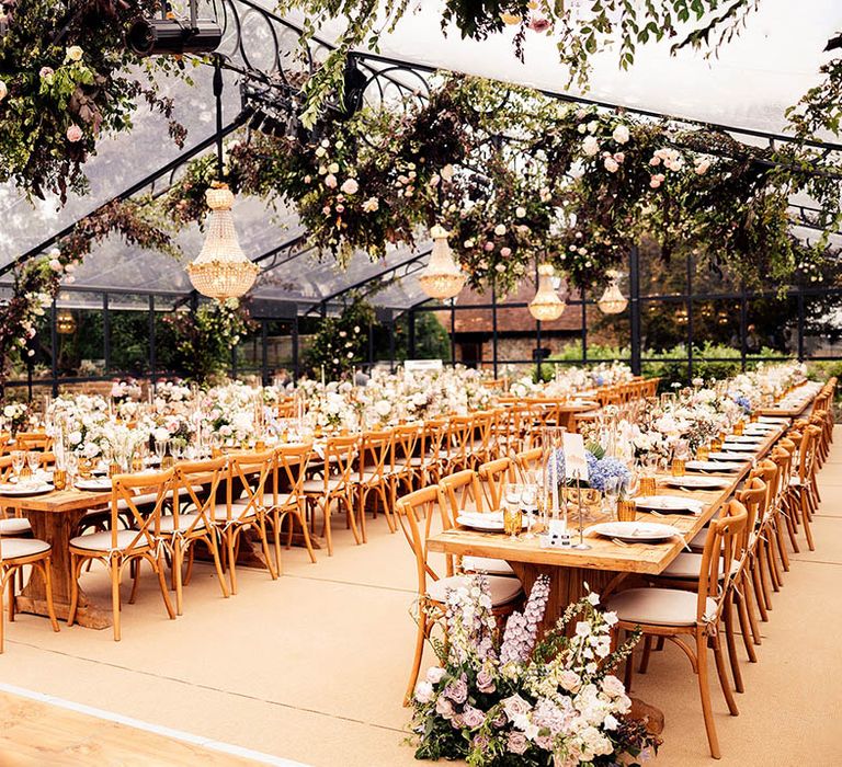 Glass marquee with long wooden banquet tables and chairs, pink and white pretty flowers with chandelier installations