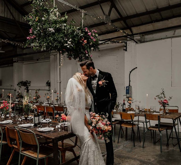 Bride in boho two piece lace bridal outfit with large mixed colourful bouquet and groom in black tux leaning on table at Hackney Studios