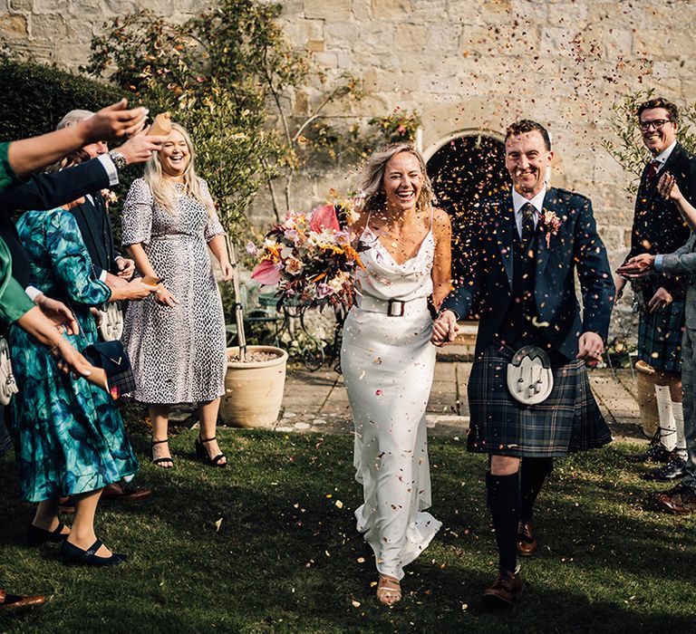 Bride in cowl neck silk slip wedding dress walks alongside her groom in kilt through colourful confetti 