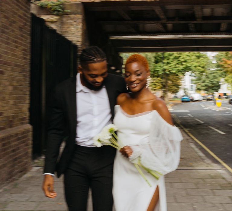 Groom in skinny black suit and bride in off shoulder wedding dress with lace puff sleeves and leg slit holding a bunch of white dahlias