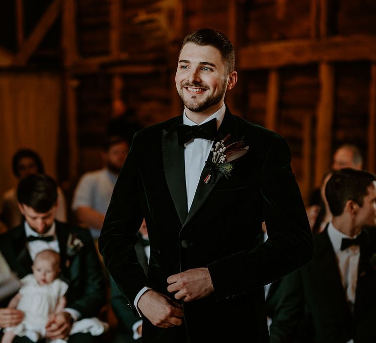 Groom wearing a green velvet tuxedo with a dried flower buttonhole waiting at the aisle for the bride 