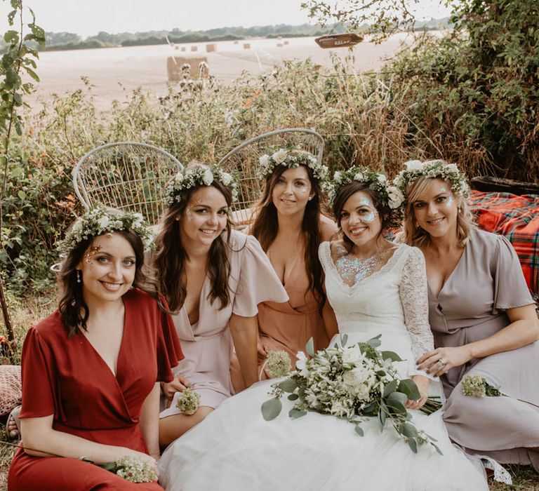 The bride sits down with her bridesmaids all wearing flower crowns and mismatched bridesmaid dresses 