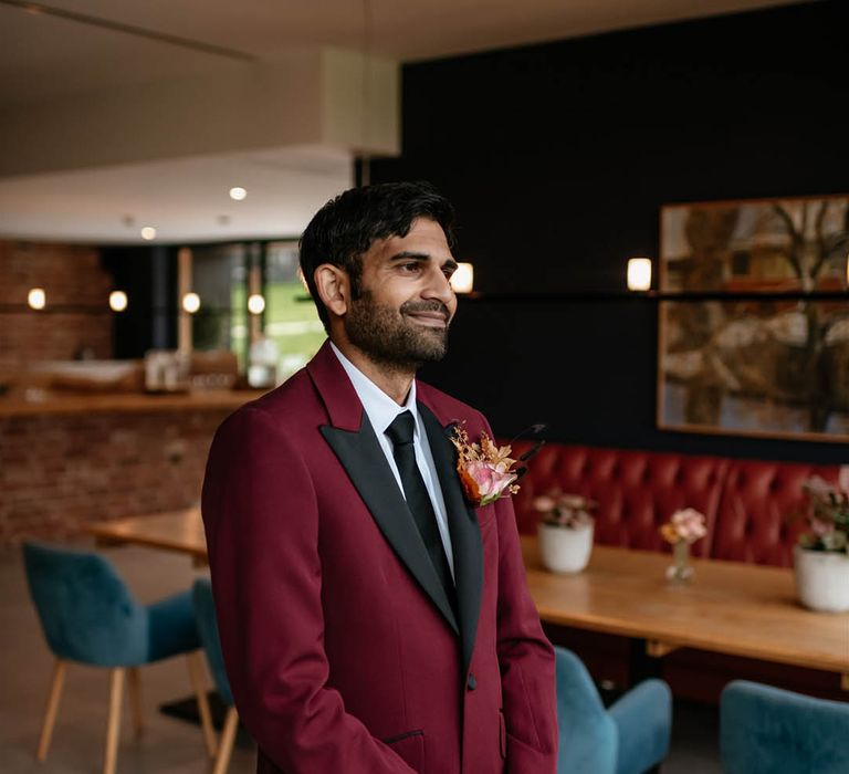 Groom stands waiting for the private first look moment in a burgundy suit with black lapels and tie with a large rose buttonhole 