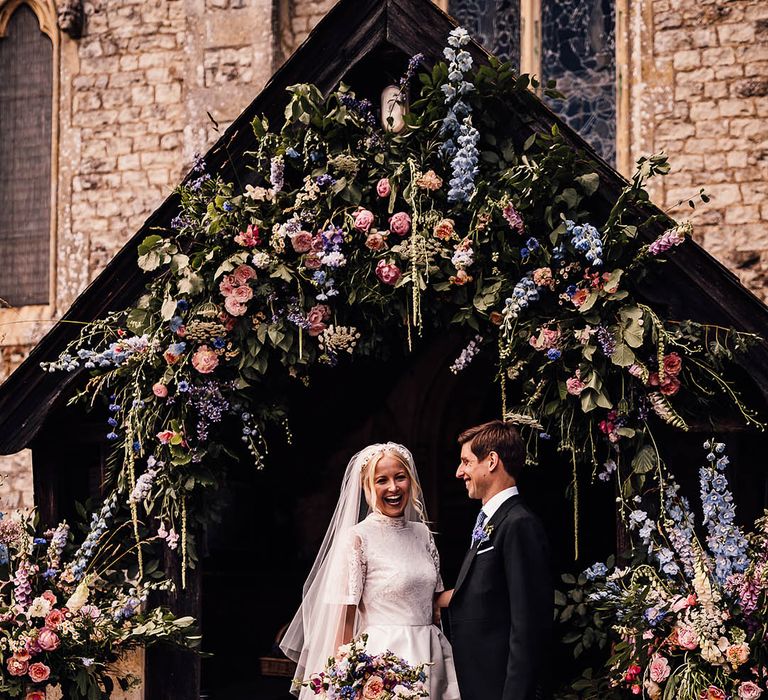 Summery pink and blue wedding flower arch and milk churns decorate the entrance to the church