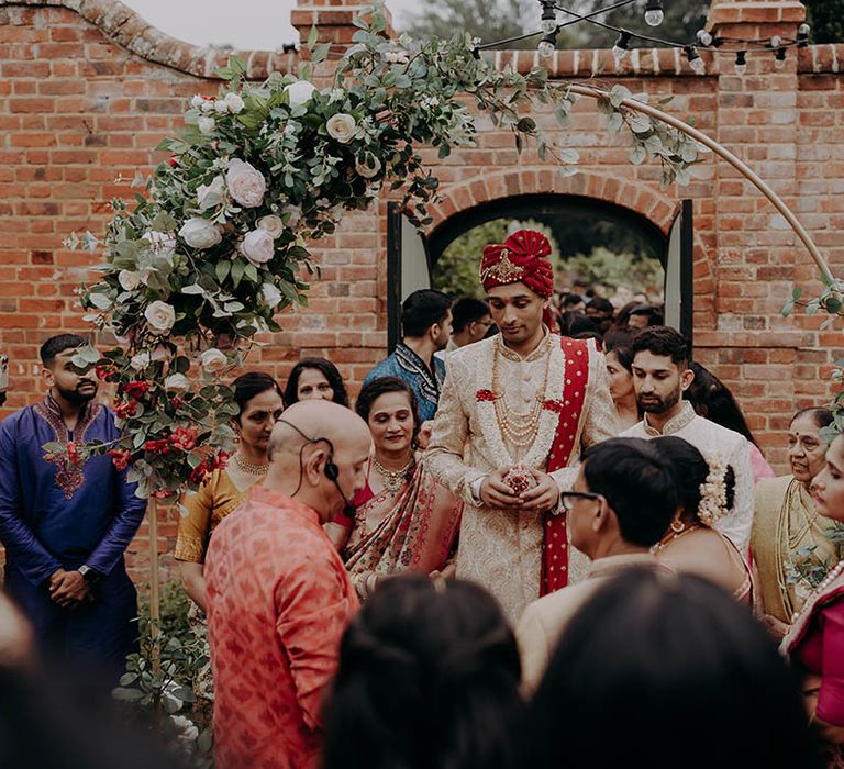 Groom wears red and gold outfit whilst stood outdoors surrounded by wedding guests 