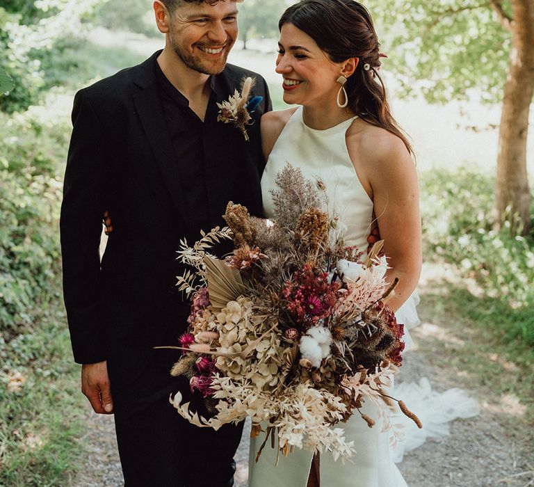 Groom in all black suit with dried bunny grass buttonhole smiling with the bride in a high neck wedding dress with DIY flower bouquet 