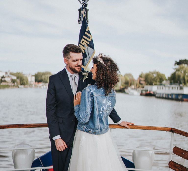 Bride wears personalised denim jacket and looks lovingly toward her groom during Thames boat ride 