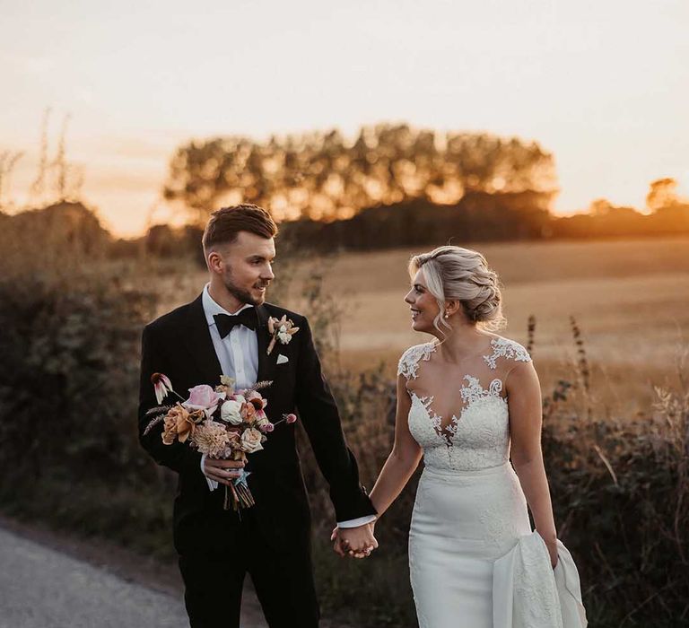 Bride in lace wedding dress with sheer, lace top and clusters of lace flowers on the shoulders and plunge neckline and groom in classic Ralph Lauren black tuxedo with black bowtie and blush rose boutonniere holding bouquet with pink garden roses, peonies, dahlias, dried flower, Persian buttercups and foliage 