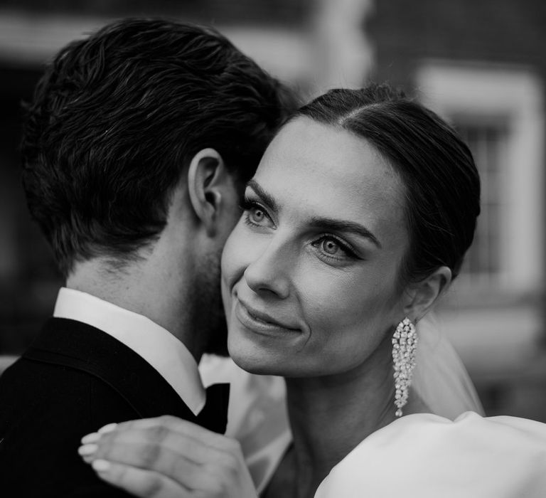 Bride in diamond drop earrings and wedding dress with puff sleeves hugging groom in black tux 