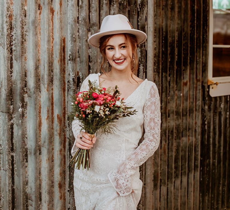 Bride in boho long sleeve lace wedding dress and white bridal fedora holding hot pink rose, peony and dried-flower bouquet 