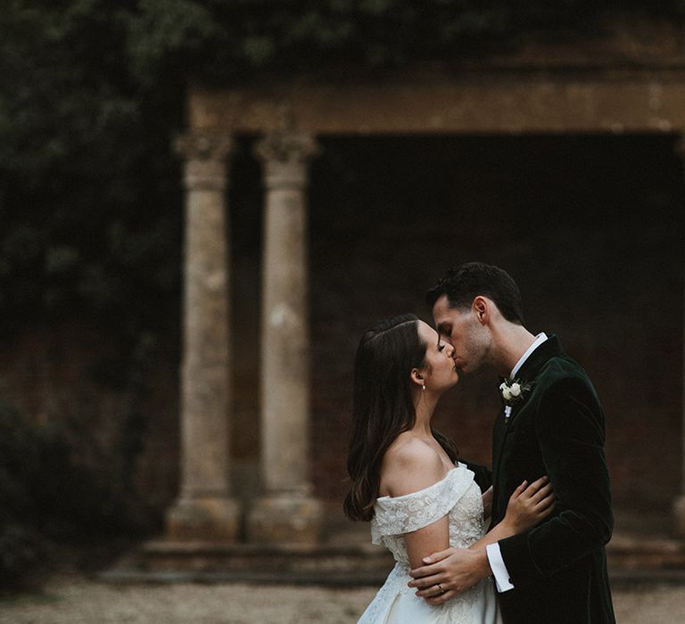 Bride in off the shoulder wedding dress kisses the groom in a dark green tuxedo 