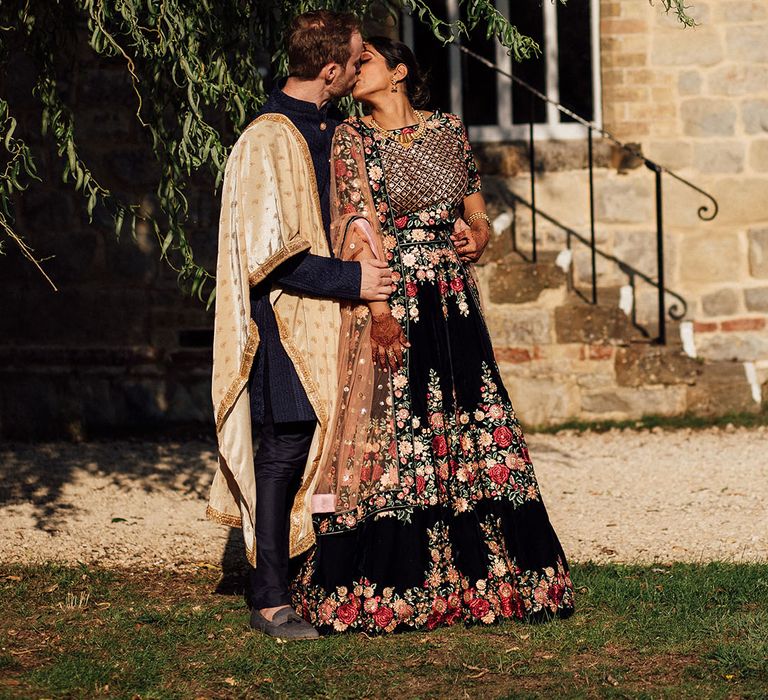 Groom wearing golden sash kisses his bride wearing a floral designed lehenga