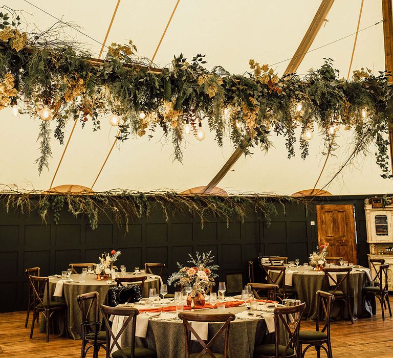 Hanging green foliage installation above round tables with green tablecloths and orange table runners 