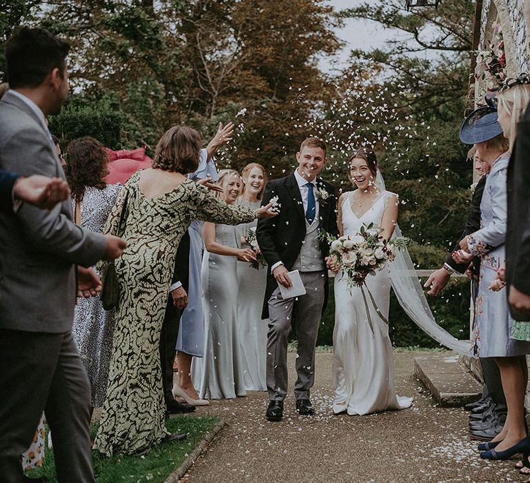 Bride & groom walk through white petal confetti outdoors after church wedding ceremony 