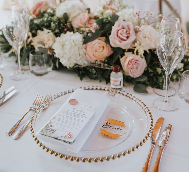 Pink and white rose table centrepiece arrangement with the bride's place setting of a clear and gold charger plate, gold cutlery, charity wedding favour with pin and pink wax seal wedding stationery 