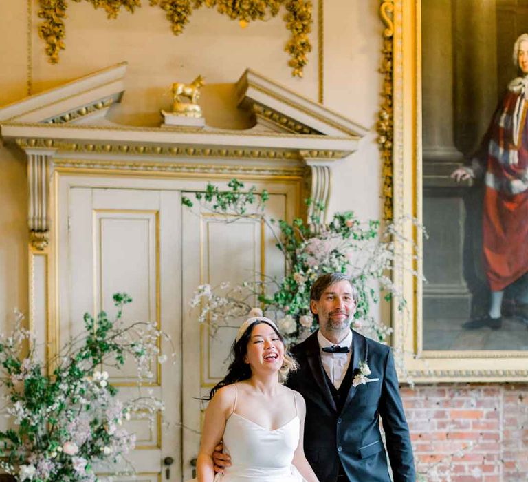 Bride in sleeveless satin wedding dress with large accent bow on the back walking with groom in classic black tuxedo, black bowtie and boutonniere by large rose, baby's-breath, foliage and dried flower floral columns holding classic spring bouquet 