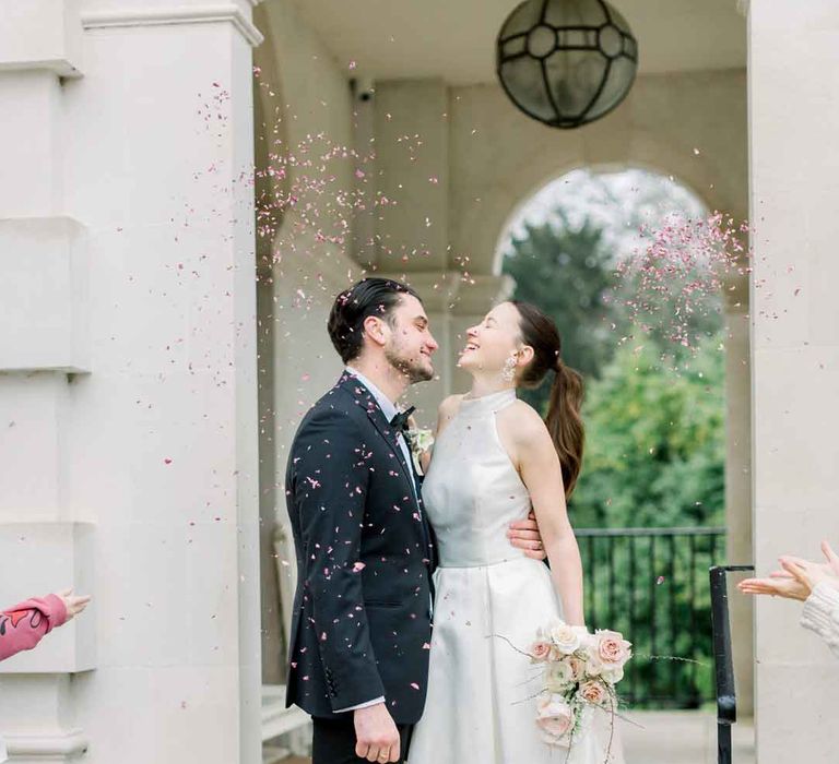 Bride in satin look halterneck wedding dress holding spring bridal bouquet standing with groom in classic black tuxedo doing petal confetti exit at wedding 