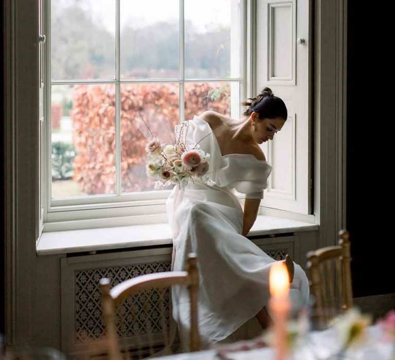 Bride in dramatic layered, sculpted off the shoulder wedding dress sitting on windowsill at St Giles House