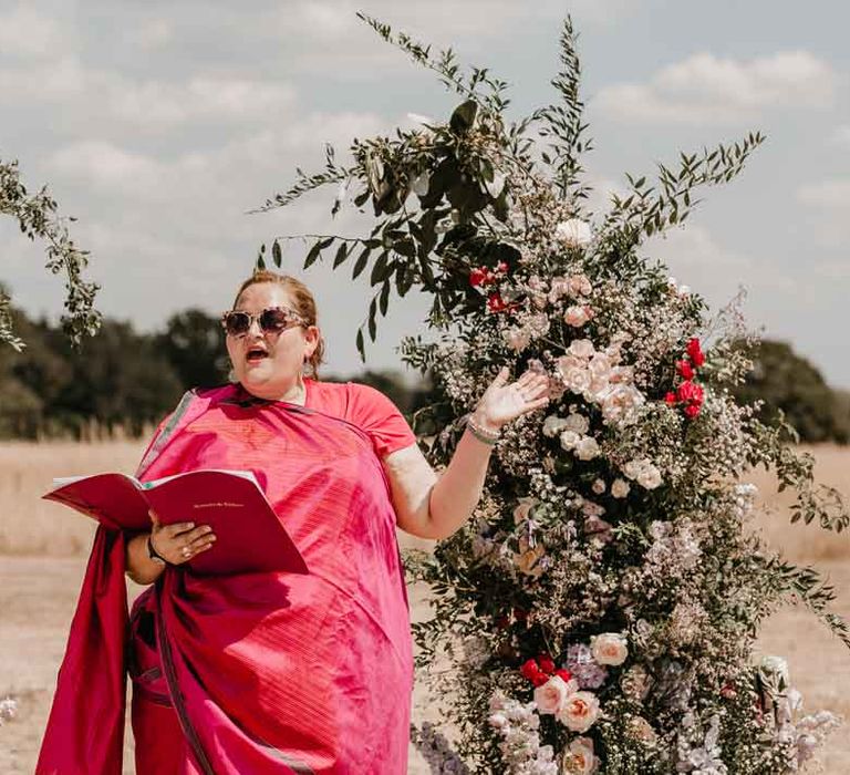 Wedding celebrant in magenta lehenga and sunglasses standing in front of large garden rose, baby's-breath and foliage floral columns