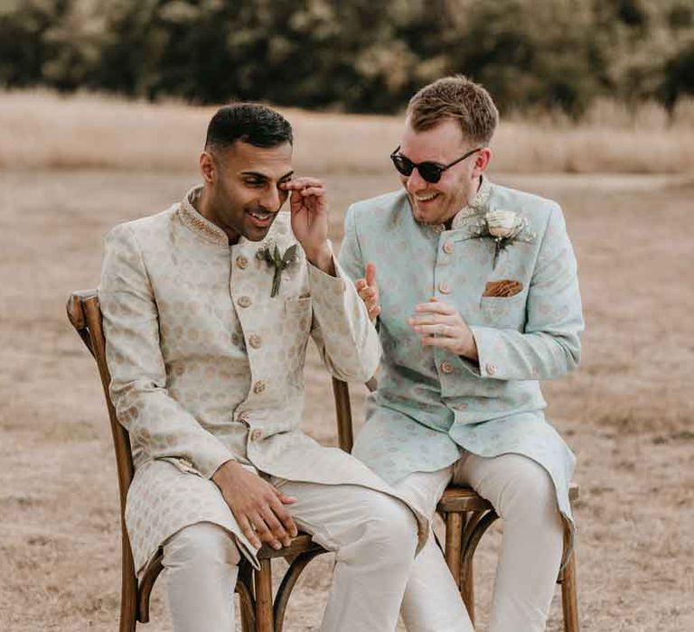 Groom in cream and gold Indian sherwani and white garden rose boutonniere sitting with groom in mint green Indian Sherwani with white garden rose and foliage boutonniere and grooms sunglasses at Gujarati wedding ceremony