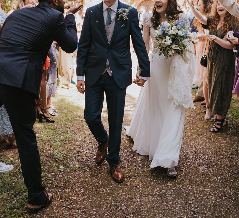 Groom in navy wedding suit with light blue tie and gray waistcoat walking with bride in lace wedding dress for confetti moment  