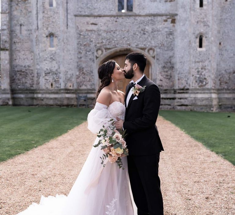 Groom in classic black tuxedo with bowtie and garden rose and eucalyptus boutonniere kissing bride in off the shoulder wedding dress with detachable puff tulle sleeves and embroidered rose 3d appliqué details holding blush garden rose and eucalyptus bridal bouquet outside of Pentney Abbey wedding venue 