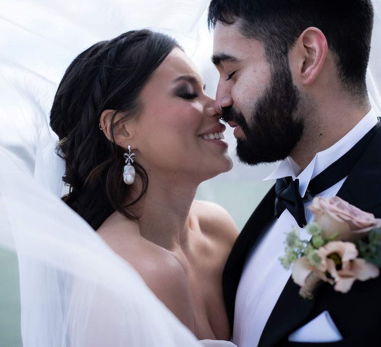 Bride in off the shoulder ethereal 3d appliqué wedding dress kissing groom in classic black tuxedo with bowtie and garden rose and eucalyptus boutonniere 