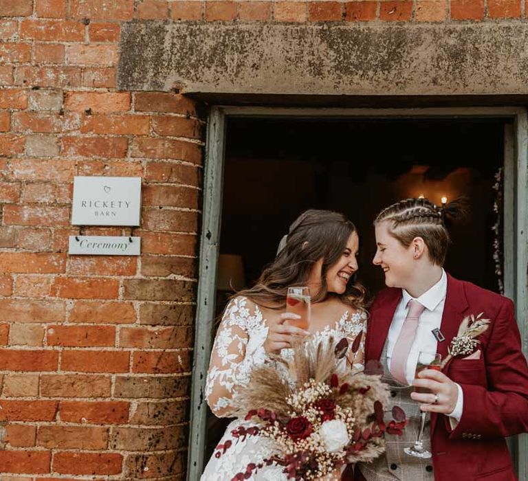 Bride in maroon bridal suit with grey waistcoat, dusky pink tie and autumnal dried flower boutonniere sipping champagne and laughing with bride in long sleeve lace illusion sleeve wedding dress with overlay holding large autumnal boho dried flower bouquet 