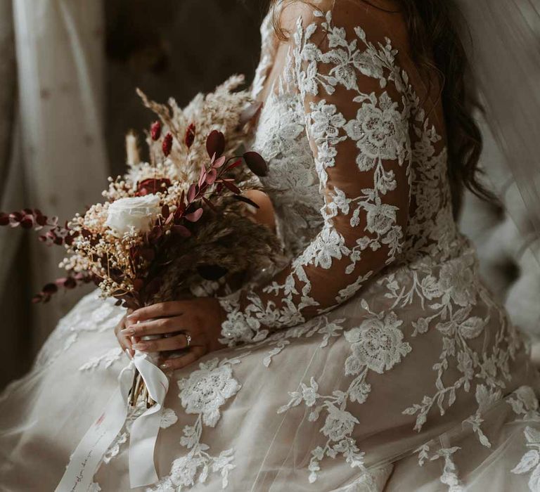 Bride wearing long sleeve lace illusion sleeve wedding dress with overlay holding boho dried flower bouquet 